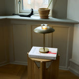 Stepp LED Outdoor Table Lamp in antique brass with a shallow round disc shade and slender stem, glowing softly atop a book on a wooden side table near a window.