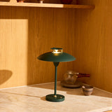 Stepp LED Outdoor Table Lamp in dark green, with a wide flat disc shade on brass posts, slim stem, and round base, shown lit on a marble surface against a wooden shelf backdrop.