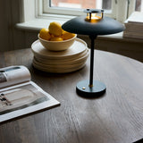 Stepp LED rechargeable table lamp in dark green with a wide flat disc shade and slim stem on a round base, shown lit on a wooden table beside a lemon bowl and stacked plates.