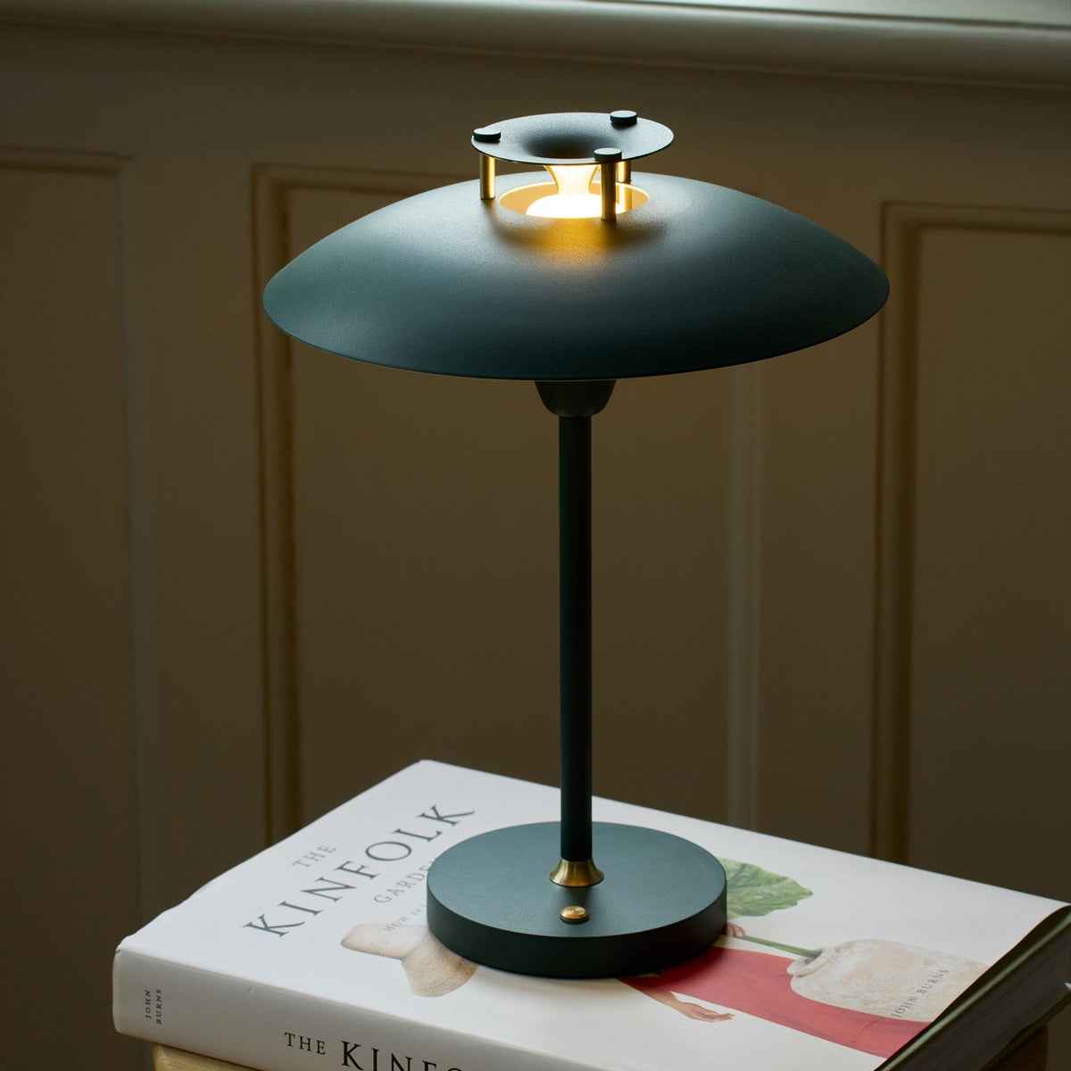Stepp LED Outdoor Table Lamp in dark green, with a wide flat disc shade held on brass posts above a stepped cap, slim stem and round base, shown lit atop a book.