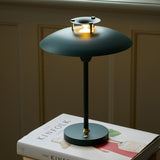 Stepp LED Outdoor Table Lamp in dark green, with a wide flat disc shade held on brass posts above a stepped cap, slim stem and round base, shown lit atop a book.