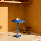 Stepp LED Outdoor Table Lamp in dark blue, wide flat disc shade with brass accents on a slim stem and round base, shown lit on a marble surface against a wood-panelled interior.