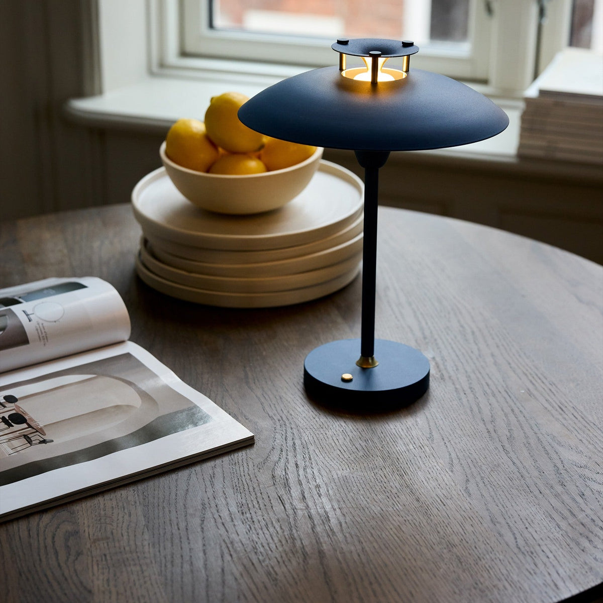 Stepp LED rechargeable table lamp in dark blue, wide flat disc shade with brass posts on a slim stem and round base, shown on a wooden table with lemons and books.