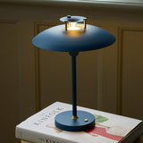 Stepp LED outdoor table lamp in dark blue, with a wide flat disc shade on brass posts glowing beneath a small stepped cap, set on a slim stem and round base atop a book indoors.