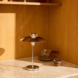 Stepp LED Outdoor Table Lamp in chrome with a wide flat disc shade and slim stem on a round base, shown on a marble counter beside a wood-panelled shelf.