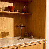 Stepp LED Outdoor Table Lamp in chrome with a wide, flat disc shade on a slim stem and round base, shown on a marble counter beside decorative bowls and vases.