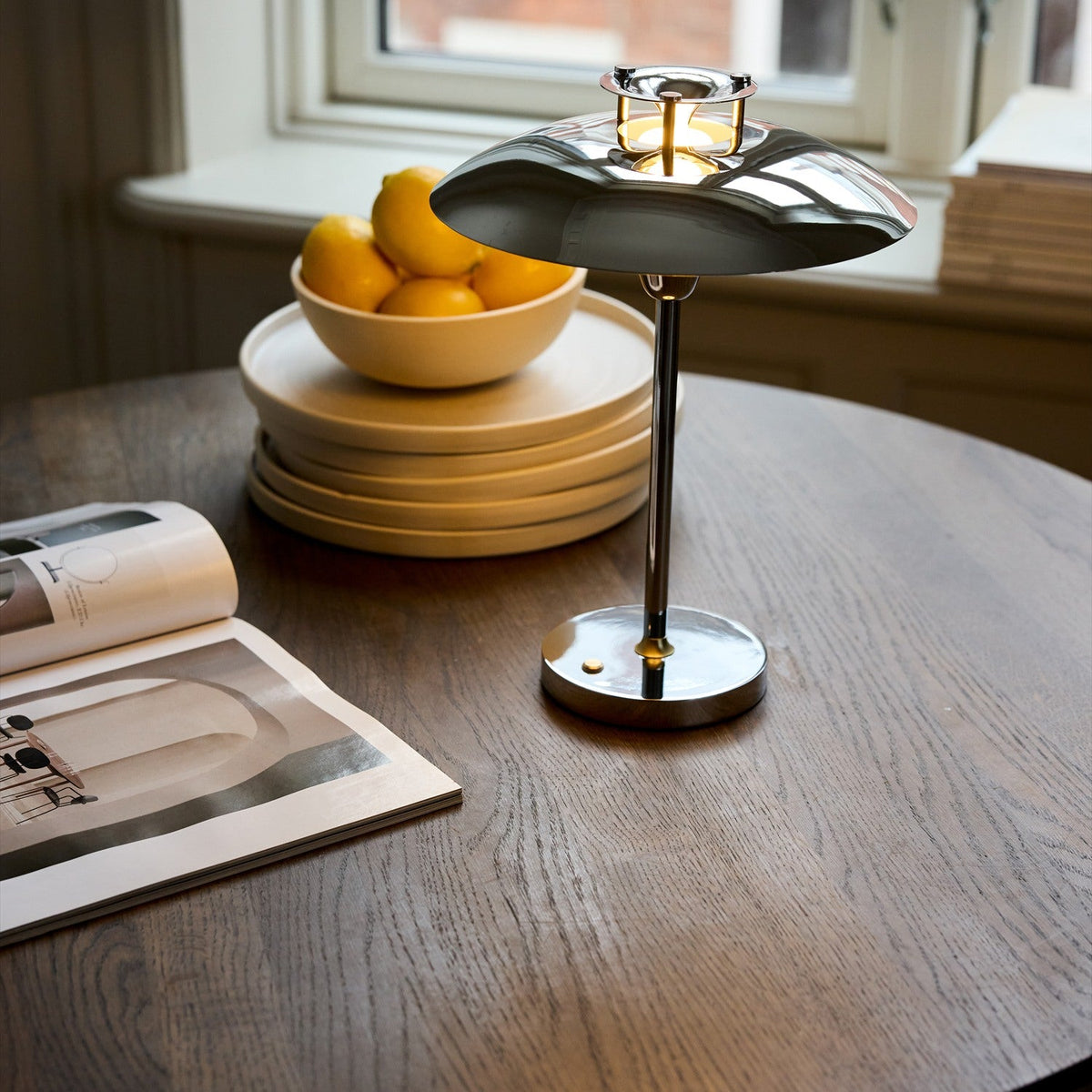 Stepp LED Outdoor Table Lamp in chrome with a wide flat disc shade and slim stem on a round base, shown lit on a wooden table beside a bowl of lemons.