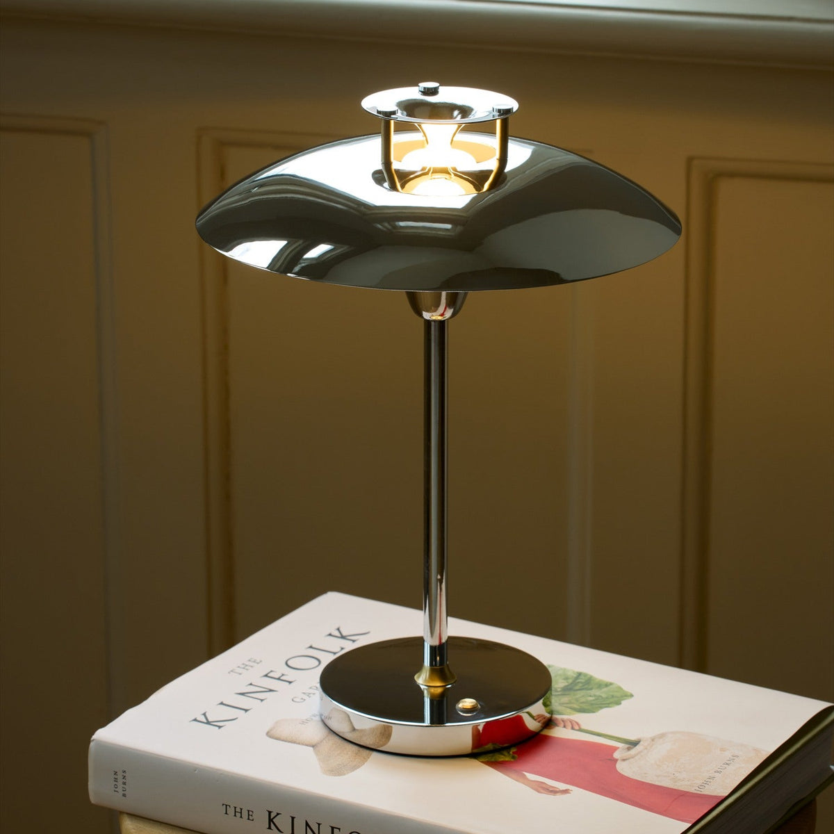 Stepp LED Outdoor Table Lamp in chrome with a wide flat disc shade, stepped cap emitting a bright ring of light, and slim stem on a round base, shown lit atop a book indoors.