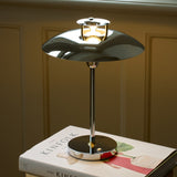 Stepp LED Outdoor Table Lamp in chrome with a wide flat disc shade, stepped cap emitting a bright ring of light, and slim stem on a round base, shown lit atop a book indoors.
