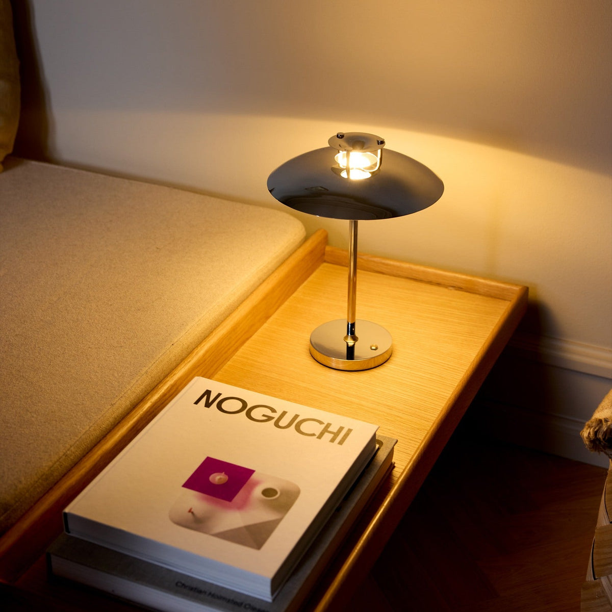 Stepp LED Outdoor Table Lamp in chrome with a wide flat disc shade and slim stem on a round base, glowing warmly on a wooden bedside tray beside books.
