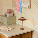 Mini Stepp LED Outdoor Table Lamp in terracotta with a wide disc shade, brass fin detail and slim stem, shown lit on a desk in a child's room setting.