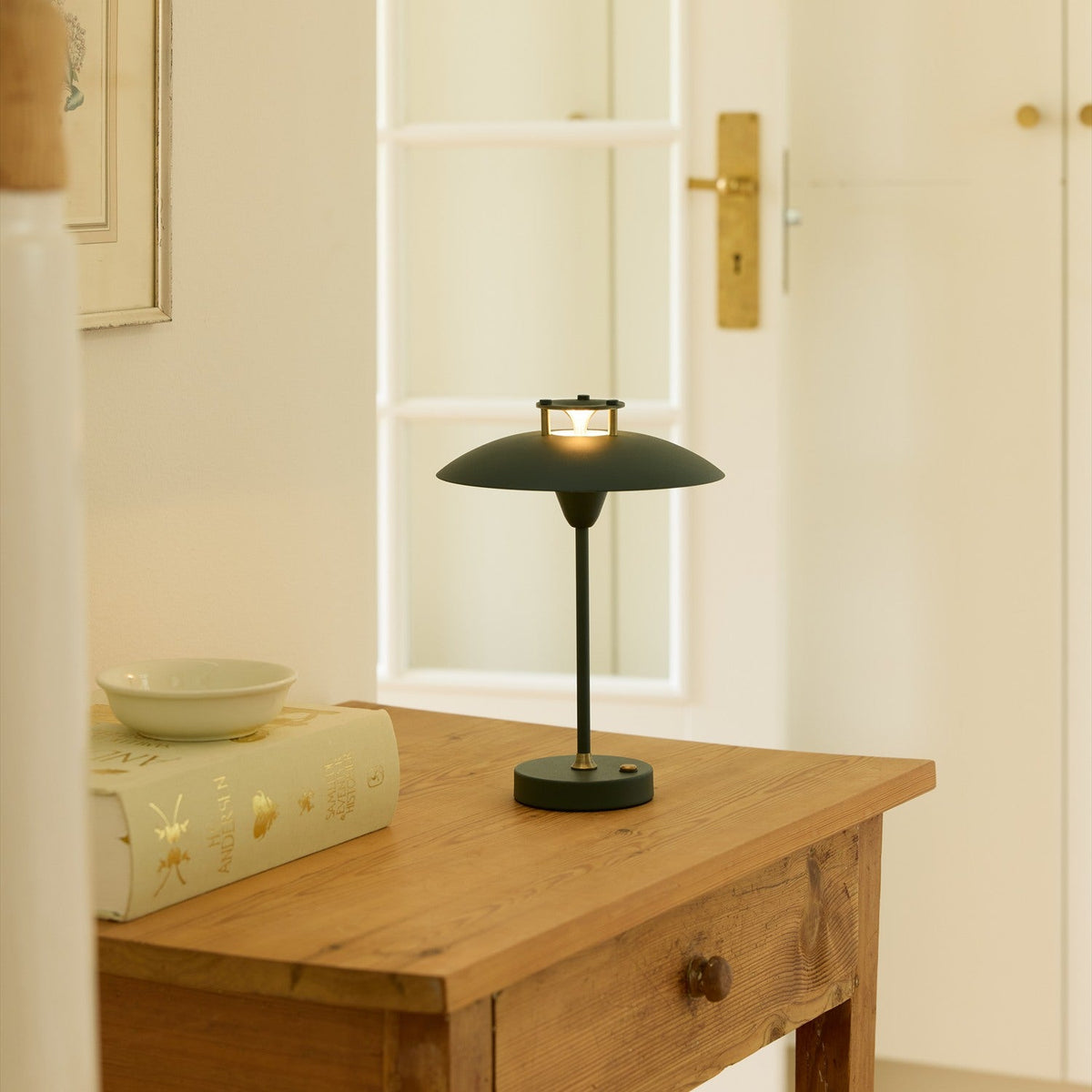 Mini Stepp LED Outdoor Table Lamp in dark green with a wide, shallow disc shade and slim stem on a round base, shown lit on a wooden console table.