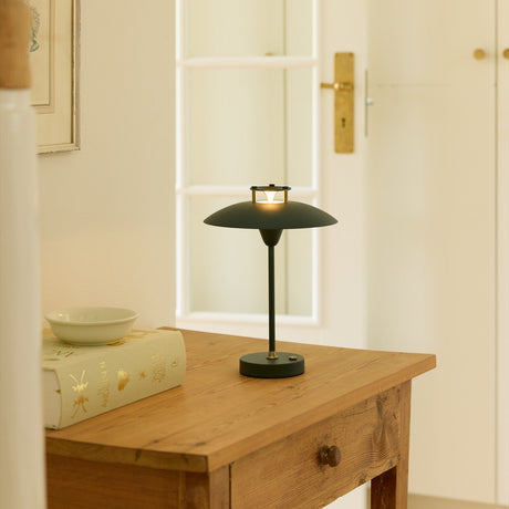 Mini Stepp LED Outdoor Table Lamp in dark green with a wide, shallow disc shade and slim stem on a round base, shown lit on a wooden console table.