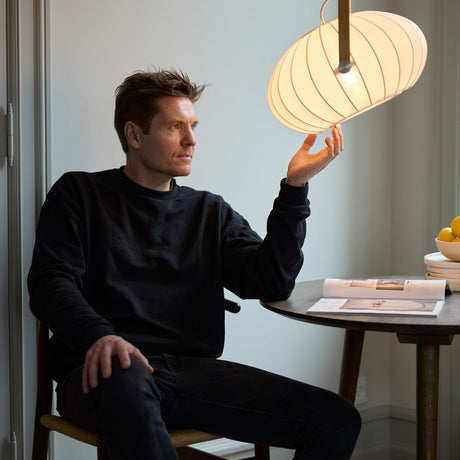 Man seated at a table reaches up to touch the DC Textile Pendant Light 40cm, a wide ribbed white fabric shade with a brass carrying-loop handle, lit above a dining setting.