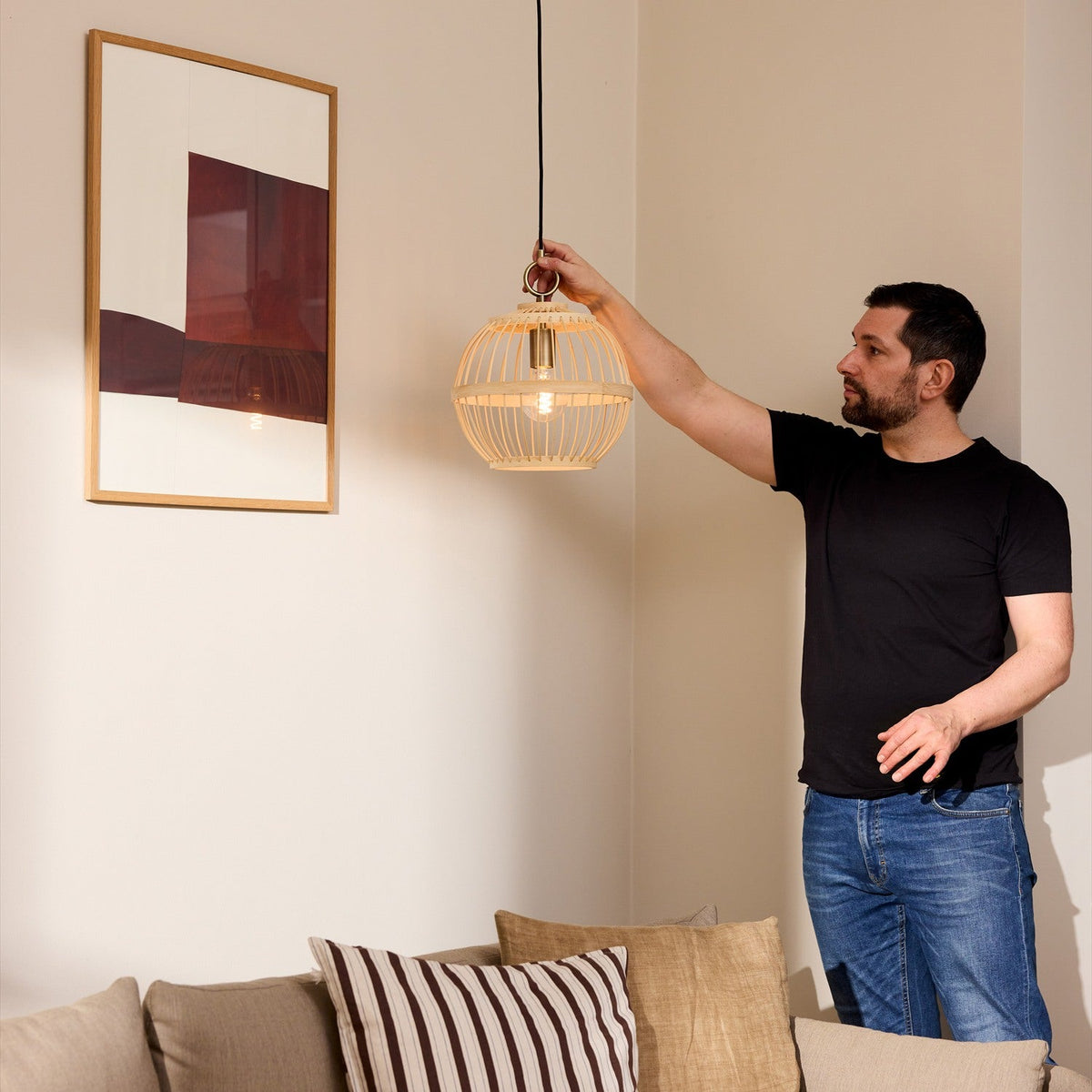 Man adjusting the Stockholm Nordic Edition Pendant Light, a round natural bamboo cage pendant with brass collar, hanging above a living room sofa.