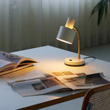 Vogue Table Lamp in cream with a cylindrical shade on a brass gooseneck and round base, glowing on a desk beside an open magazine.