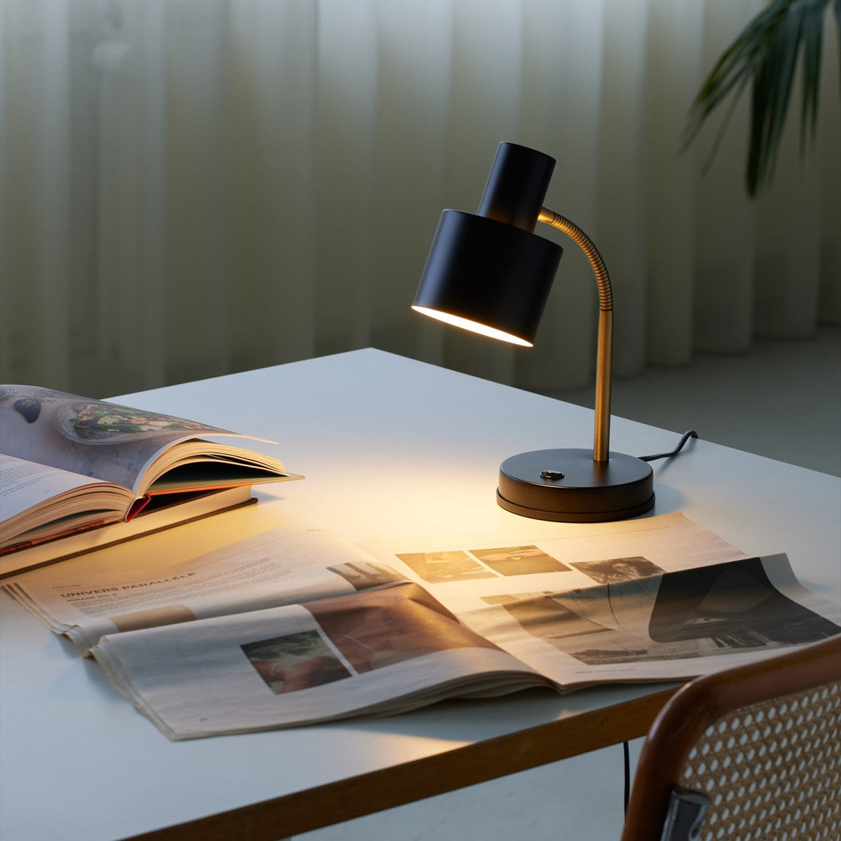 Vogue Table Lamp in black with cylindrical shade on a brass gooseneck arm and round base, lit on a desk beside an open magazine.