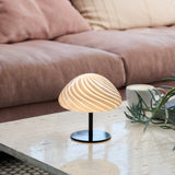 Mini Mush Glass Cable Table Lamp with a swirled light grey glass dome on a slender black metal stem and round base, glowing softly on a marble table beside a pink sofa.