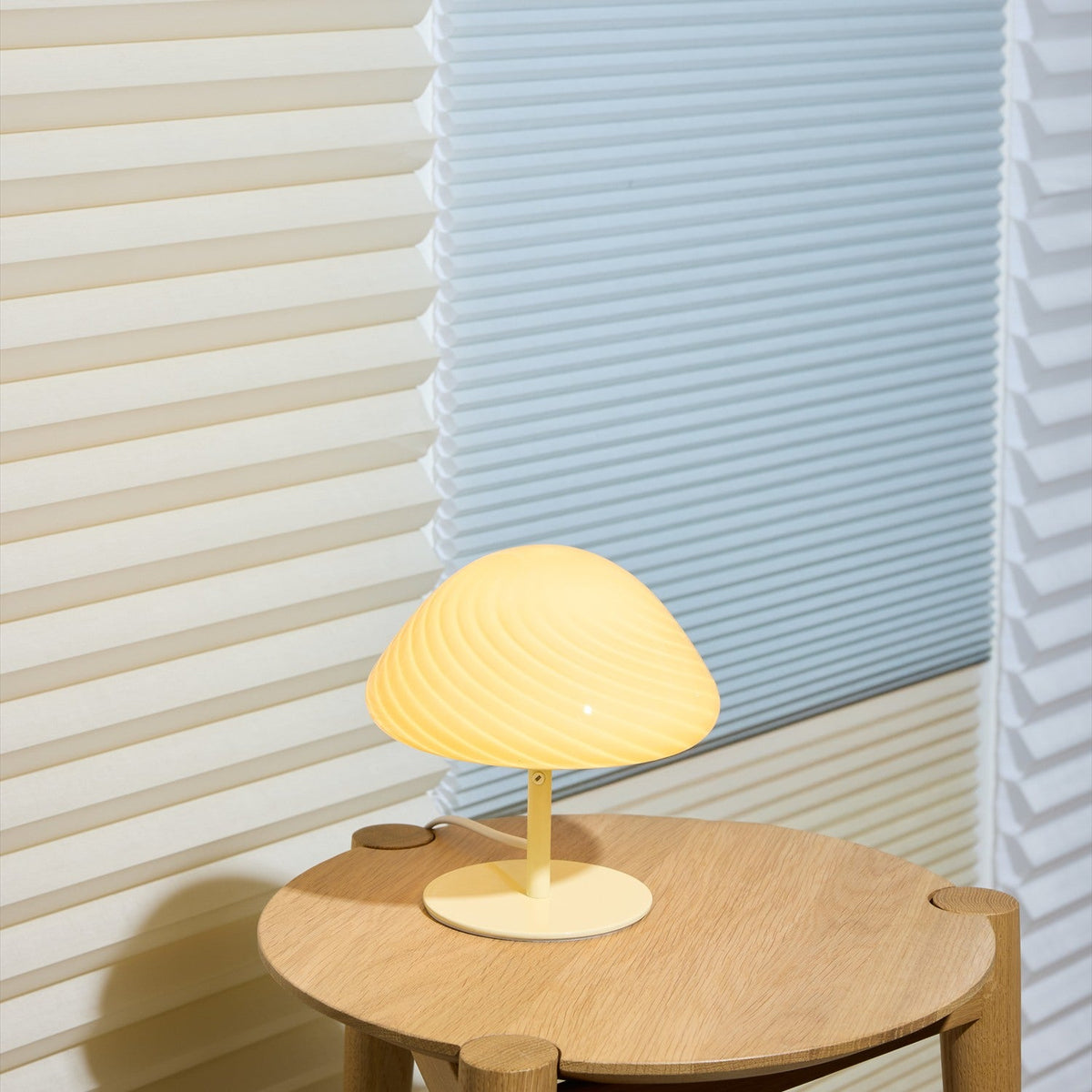 Mini Mush Glass Cable Table Lamp in cream with a spiral-fluted dome shade on a slim black stem and round base, shown lit on a round wooden side table.