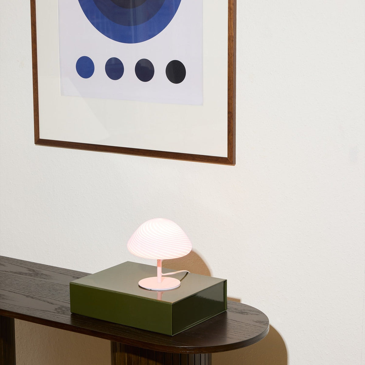 Mini Mush Glass Cable Table Lamp in rose, mushroom-shaped fluted glass shade on a slim pink metal base, shown lit atop a green box on a dark console table beneath framed wall art.