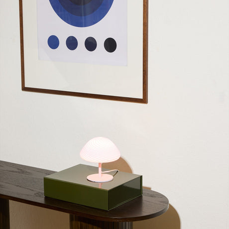 Mini Mush Glass Cable Table Lamp in rose, mushroom-shaped fluted glass shade on a slim pink metal base, shown lit atop a green box on a dark console table beneath framed wall art.