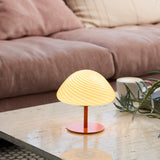 Mini Mush Glass Cable Table Lamp in orange, with a wide fluted glass dome shade glowing warm cream, set on an orange metal stem and base on a coffee table beside a pink sofa.