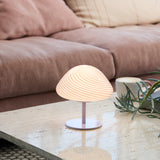 Mini Mush Glass Cable Table Lamp in purple, with a wide spiral-fluted glass dome on a slim metal stem and round base, shown lit on a coffee table beside a sofa.