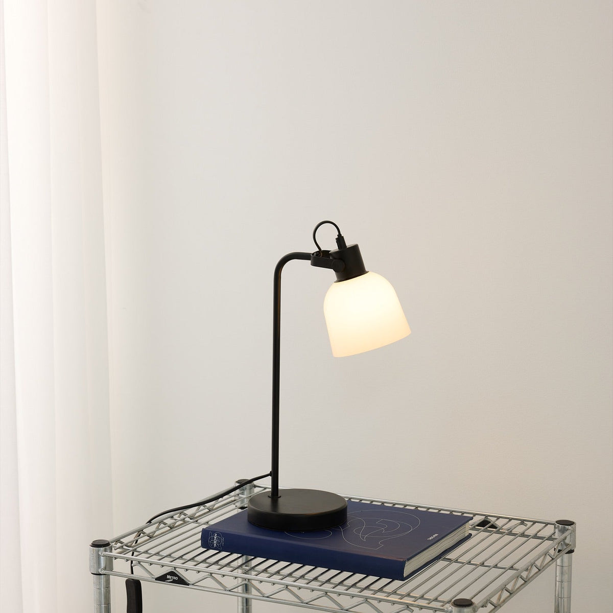 Glow Table Lamp in black and white, featuring an opal glass bell shade on a slim black metal post with round base, lit atop a book on a wire shelf against a plain wall.