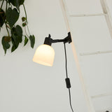 Glow Clip-On Table Lamp in black and white, with an opal glass bell shade on a jointed black arm, clamped to a white ladder rung near hanging plant leaves.