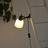 Glow Clip-On Table Lamp in black and white, with an opal glass bell shade on a jointed black arm and clamp, shown mounted beside a ladder near trailing plant leaves.