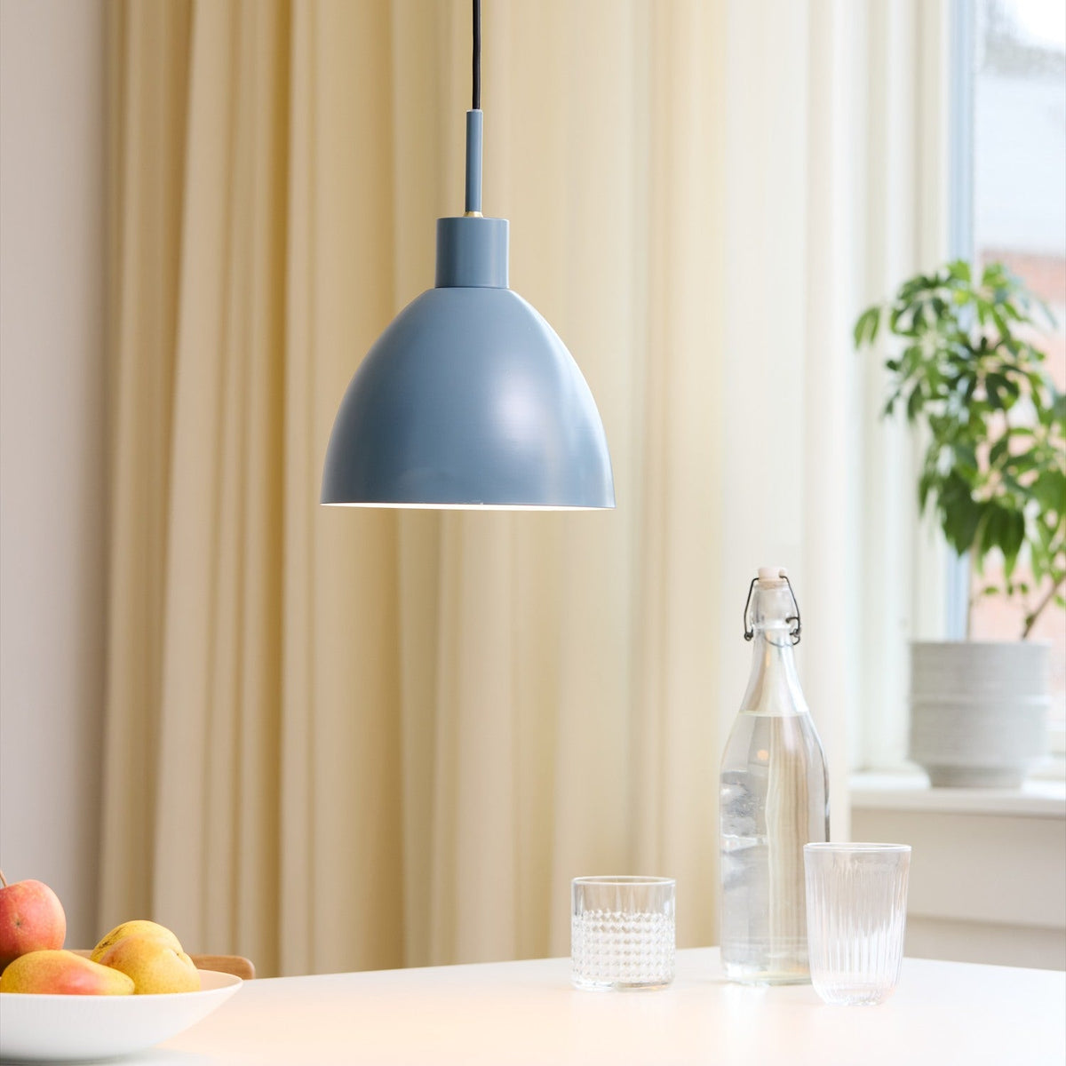 Rock Pendant Light 20cm in petrol blue, a bell-shaped metal shade with white interior, hanging above a dining table set with glasses and a bottle.