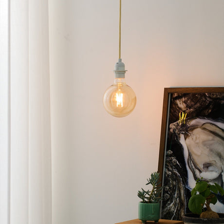 Basic Socket Textile Cable Cord Set 2m in cream, shown as a pendant light with white lampholder and amber globe bulb, hanging near a curtain in a living space.