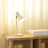 Style Table Lamp in cream and antique brass, angled cone shade on a brass knuckle joint and slim post with round base, shown lit on a wooden desk beside books.