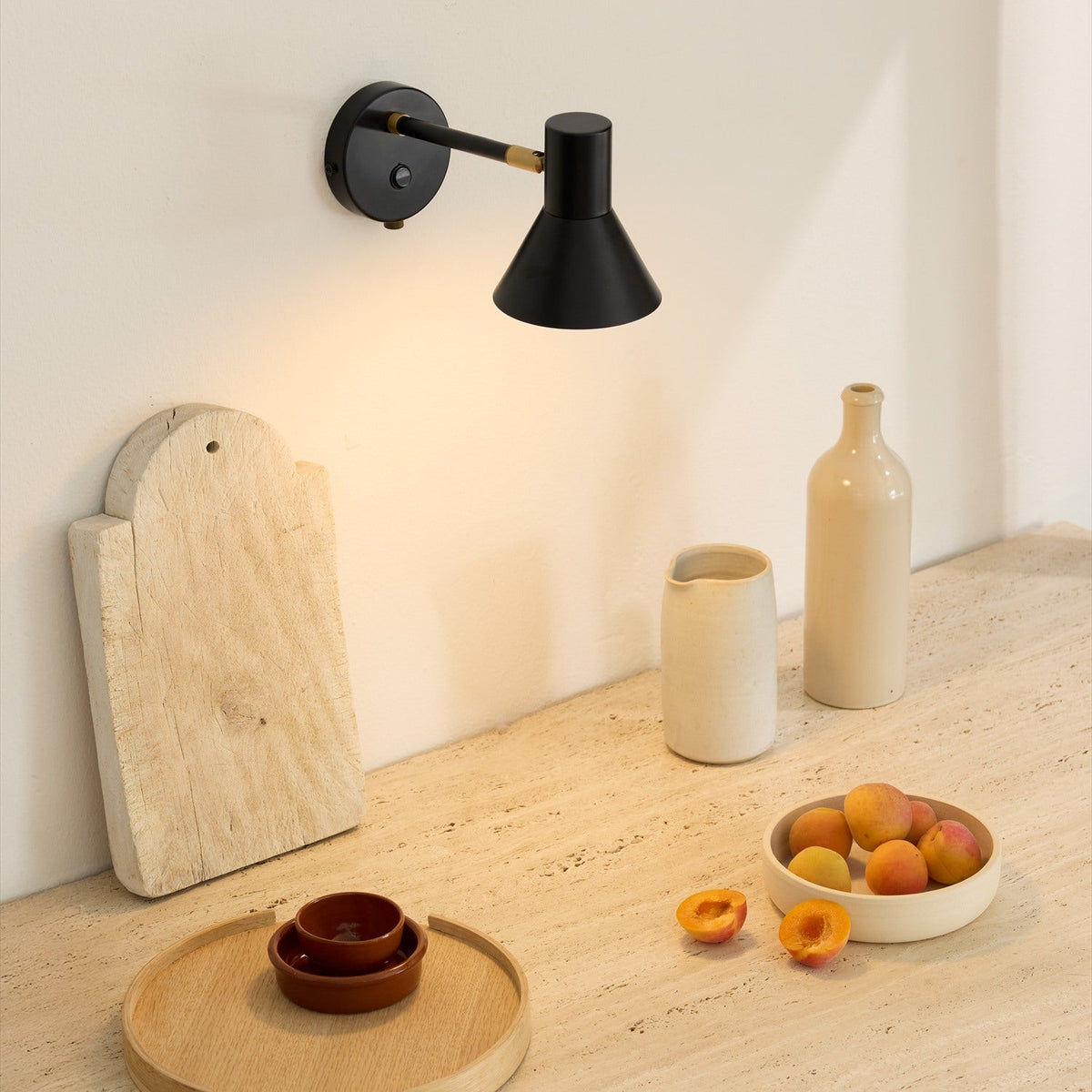 Style wall light in black and antique brass with a downward-facing cone shade, mounted above a kitchen counter styled with ceramic vases and fruit.