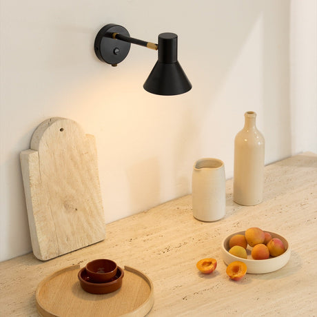 Style wall light in black and antique brass with a downward-facing cone shade, mounted above a kitchen counter styled with ceramic vases and fruit.