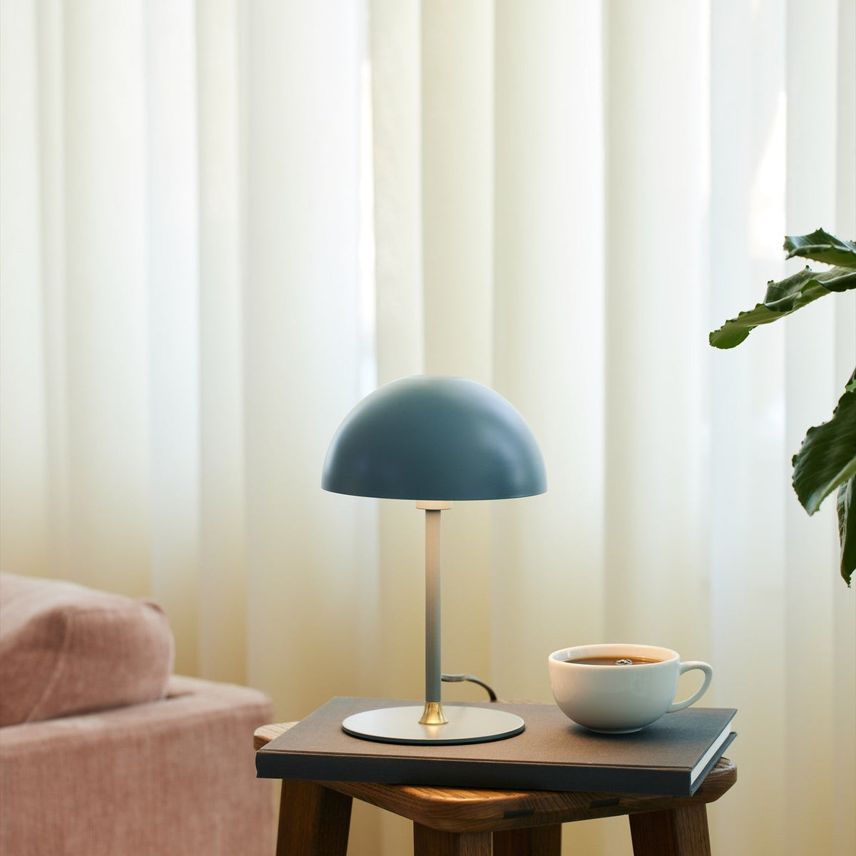 Boogie Table Lamp 16cm in petrol blue with dome shade and brass stem detail, styled on a side table beside a coffee cup in a living room setting.
