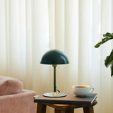 Boogie Table Lamp 16cm in dark green with a dome shade and brass base accent, shown on a side table beside a coffee cup in a living room setting.