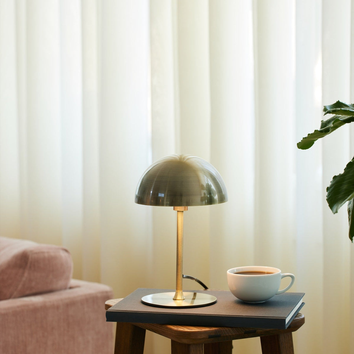 Boogie Table Lamp 16cm in antique brass with dome shade and slim stem on round base, styled on a side table beside a coffee cup in a living room.