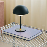 Moon LED Table Lamp in dark green with a dome shade and round base, shown on a book atop a wire shelving unit in a living room setting.