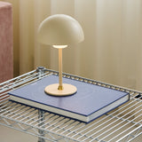 Moon LED Table Lamp in cream with dome shade and brass base detail, shown lit atop a blue book on a wire shelving unit.