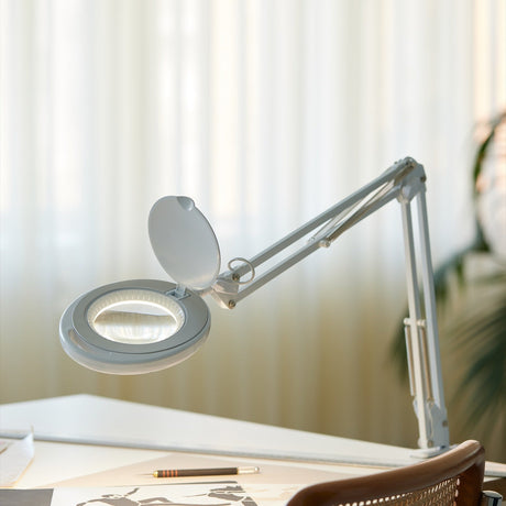 Magni LED Archi Clip Desk Table Lamp in white, with a round magnifying lens head, hinged cover, and folding sprung arm, shown clamped on a desk beside a pen and paper.