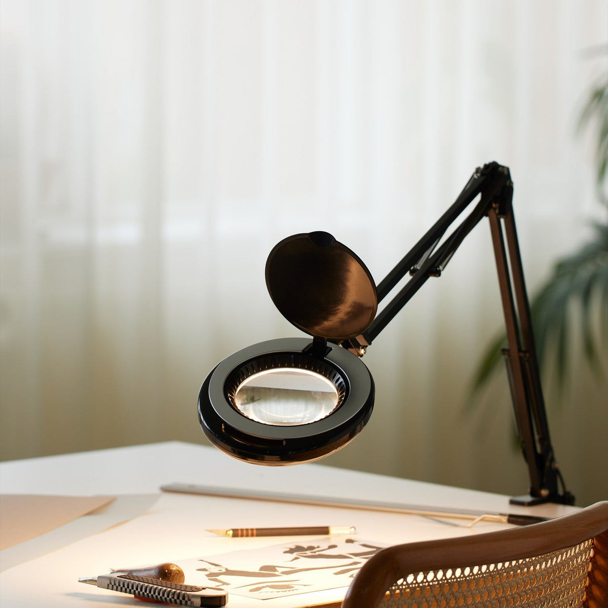Magni LED Archi Clip Desk Table Lamp in black, with a round magnifying head on a folding sprung arm, shown clamped to a desk in a home office setting.