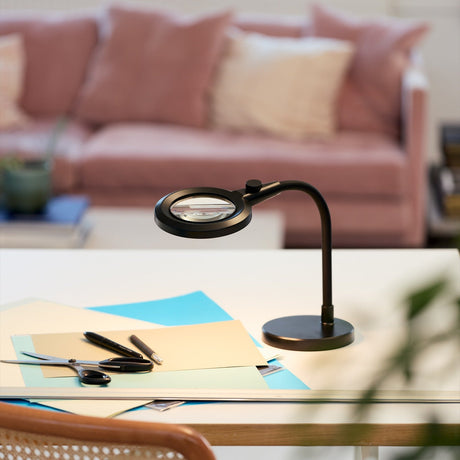 Magni Portable Superflex Charge Table Lamp in black, with a round magnifying lens head on a flexible gooseneck arm and round base, shown on a desk with stationery and a pink sofa in the background.