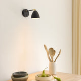Room wall light in black and brass, cone shade angled downward on a black arm with brass knuckle joints, mounted above a wooden table with pears, bowls and wooden utensils.