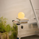 Candy Table Lamp 20cm in cream, a spiral-fluted glass globe glowing warm on a wooden block atop a metal storage case, styled with a plant and ladder nearby.