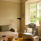 Stepp Floor Lamp in black and antique brass, with a wide flat shade and slim stem on a round base, shown beside a striped armchair in a living room.