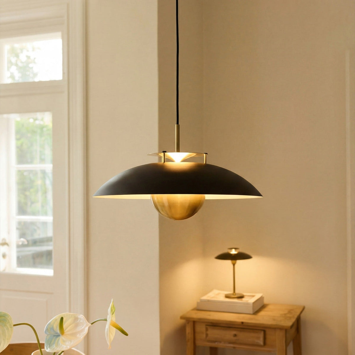 Stepp LED Pendant Light 46cm in black and antique brass, wide flat disc shade with brass fins and hanging brass ball, shown lit above a wooden side table in a living room.