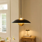Stepp LED Pendant Light 46cm in black and antique brass, wide flat disc shade with brass fins and hanging brass ball, shown lit above a wooden side table in a living room.