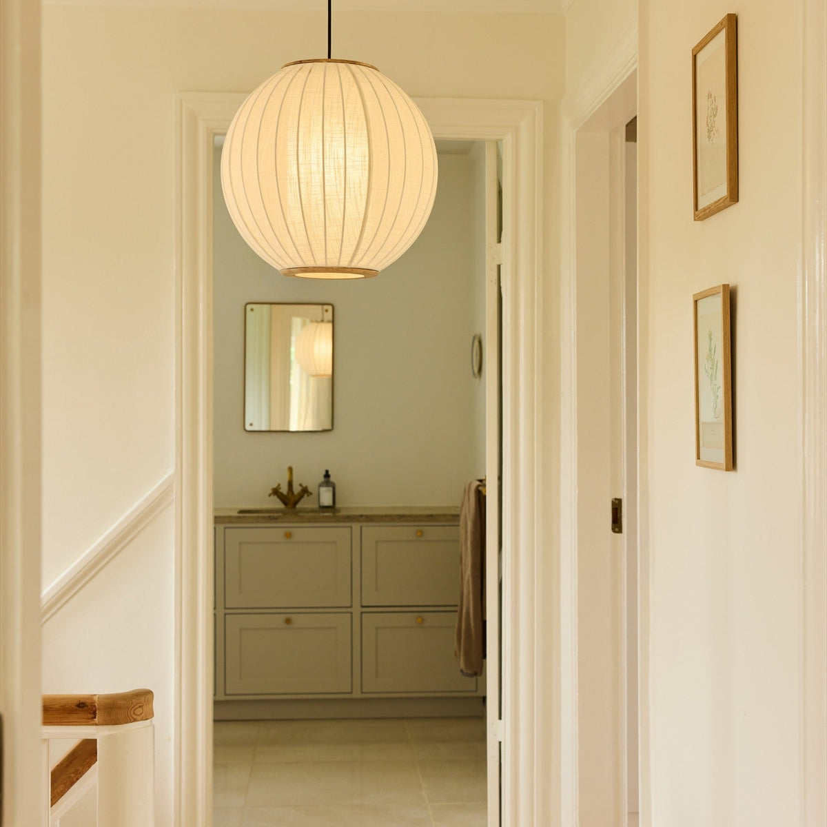 Balloon Pendant Light 40cm in white and beige, a pleated fabric globe with brass top and bottom rings, hung in a hallway leading to a bathroom with framed art nearby.