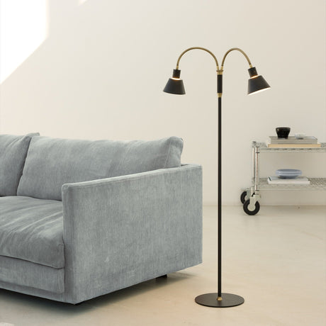 Space LED 2F Floor Lamp in black and antique brass, with two curved goosenecks and cone shades on a slim stem, shown beside a grey sofa.